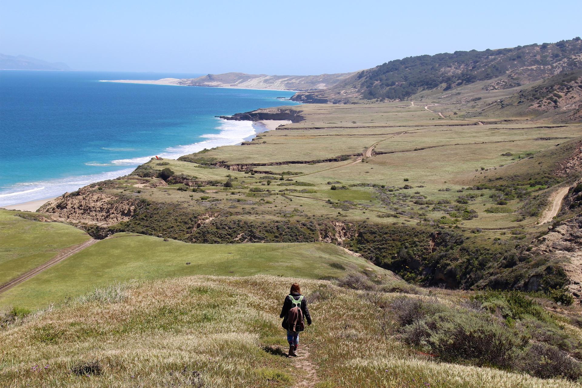 Hiker on a dirt path overlooking a vast sunny coastline and green hills.