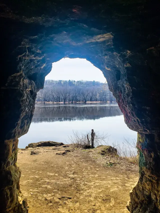 View from a dark cave opening overlooking a river and distant bare trees.