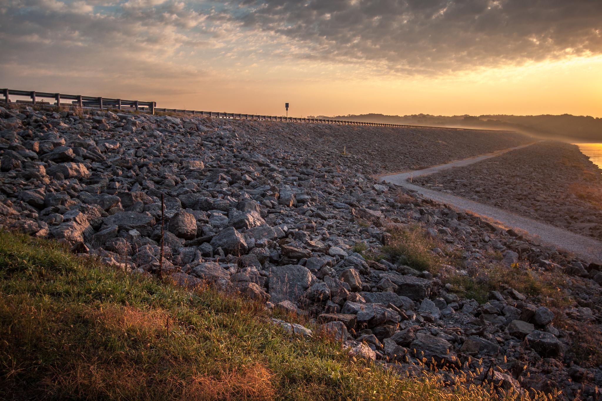 Rocky embankment with a road and path at sunset, person walking in distance.