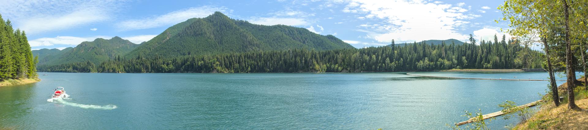 Panoramic lake view with forested mountains, bright sun, and a small boat.
