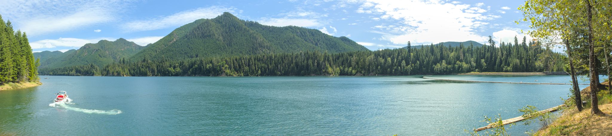 Panoramic lake view with forested mountains, bright sun, and a small boat. - Wynoochee Lake Shore Trail