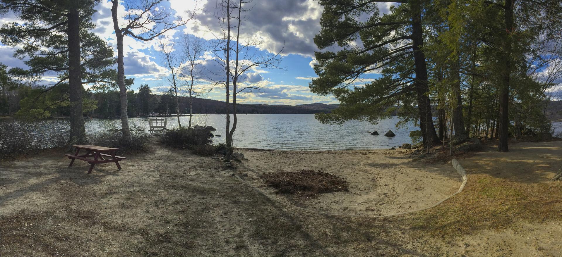 Sandy lake shore with trees, a picnic table, and distant hills under clouds.