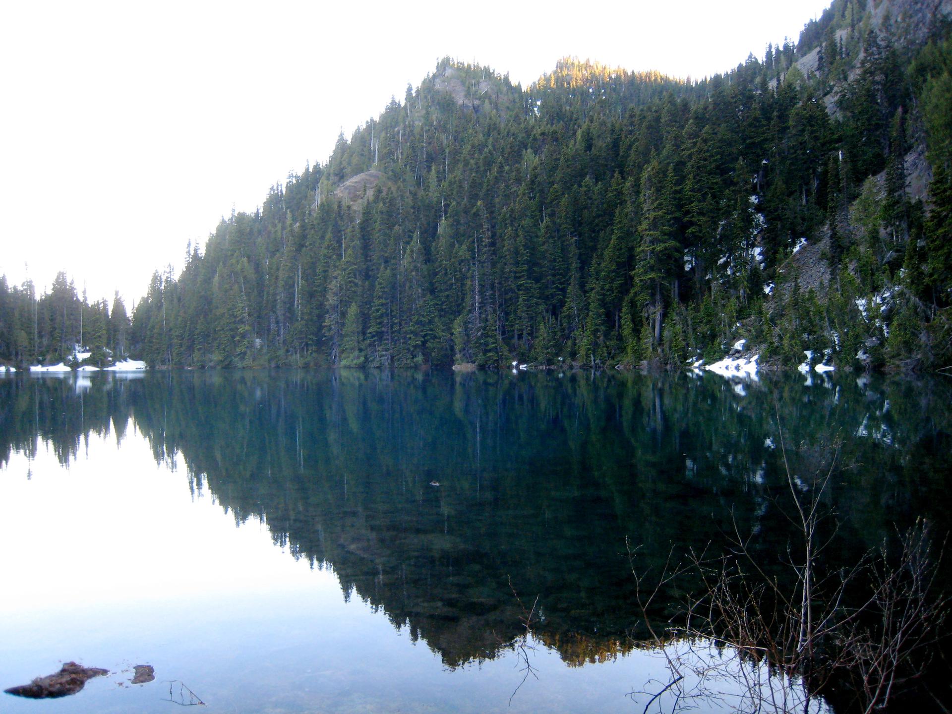 Calm mountain lake reflects pine-covered slopes and a bright, clear sky.