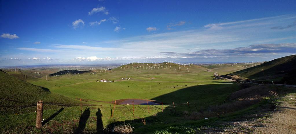 Panoramic view of green valleys with distant wind turbines under a blue sky.