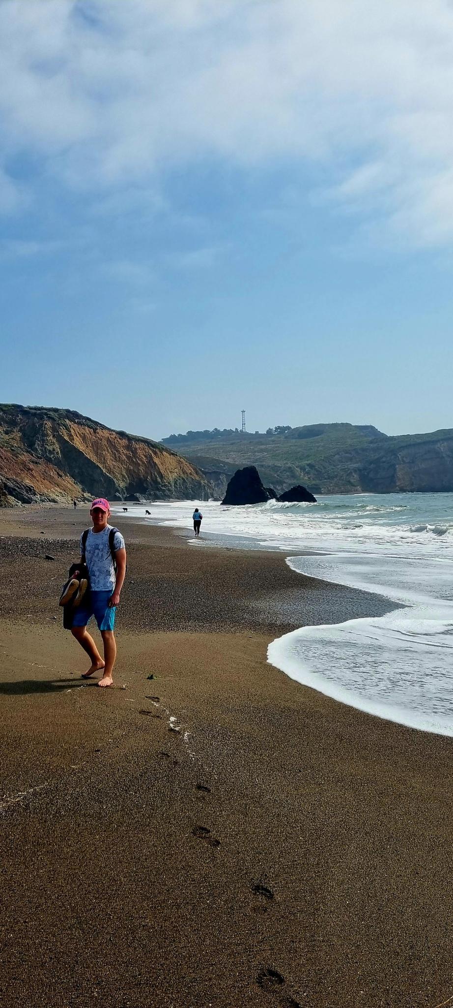 Man with bag walking on a dark sand beach with waves and cliffs.
