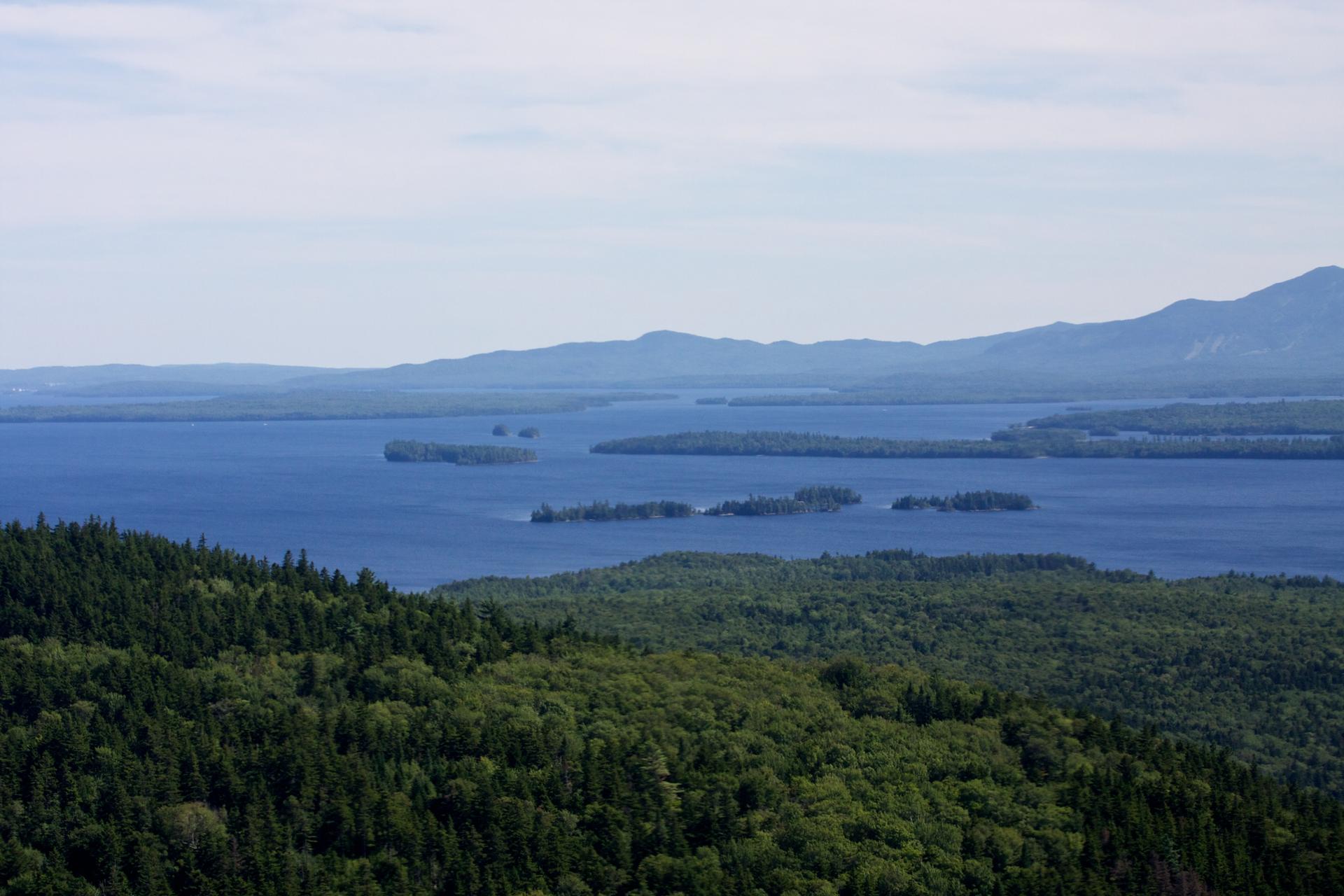 Expansive blue lake with islands, surrounded by lush green forests and distant mountains.