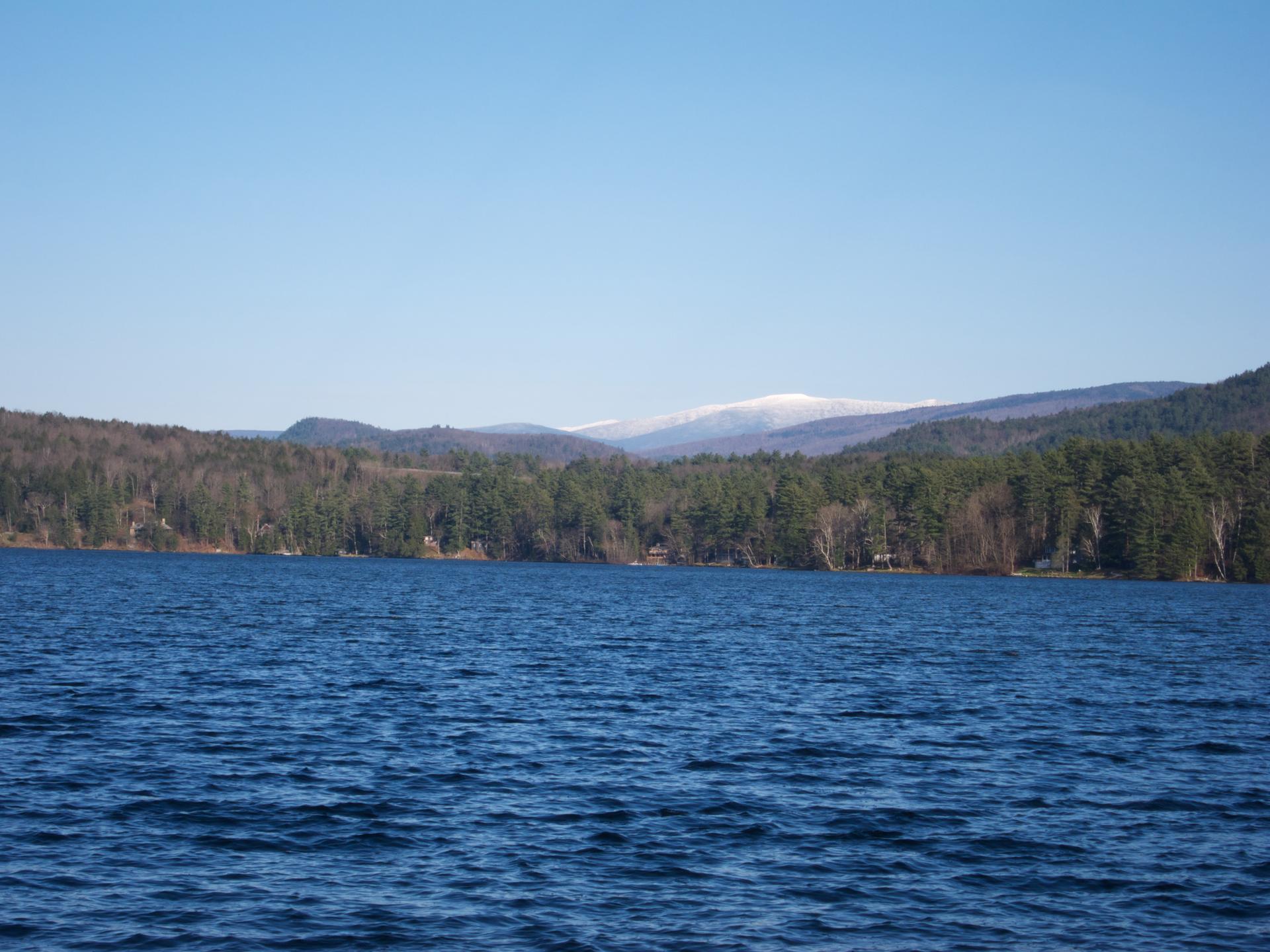 Blue lake with forested shore, distant snow-capped mountains under clear sky.