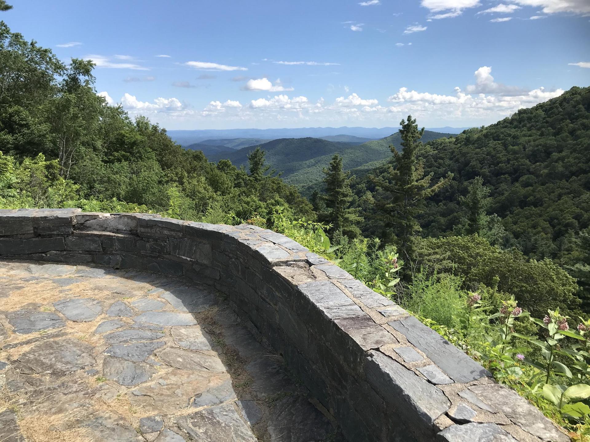 A stone overlook provides a sunny view of lush green mountain ranges.