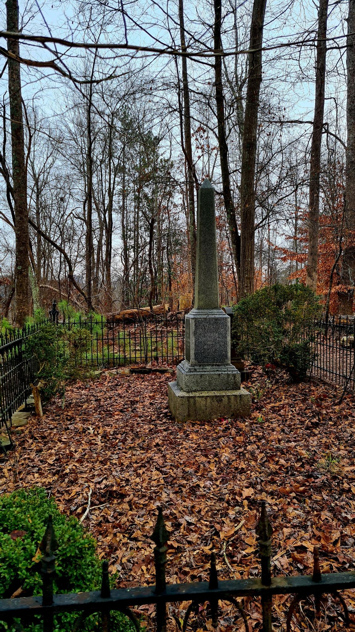 Dark obelisk monument in a fenced, leaf-covered forest clearing on a dim day.