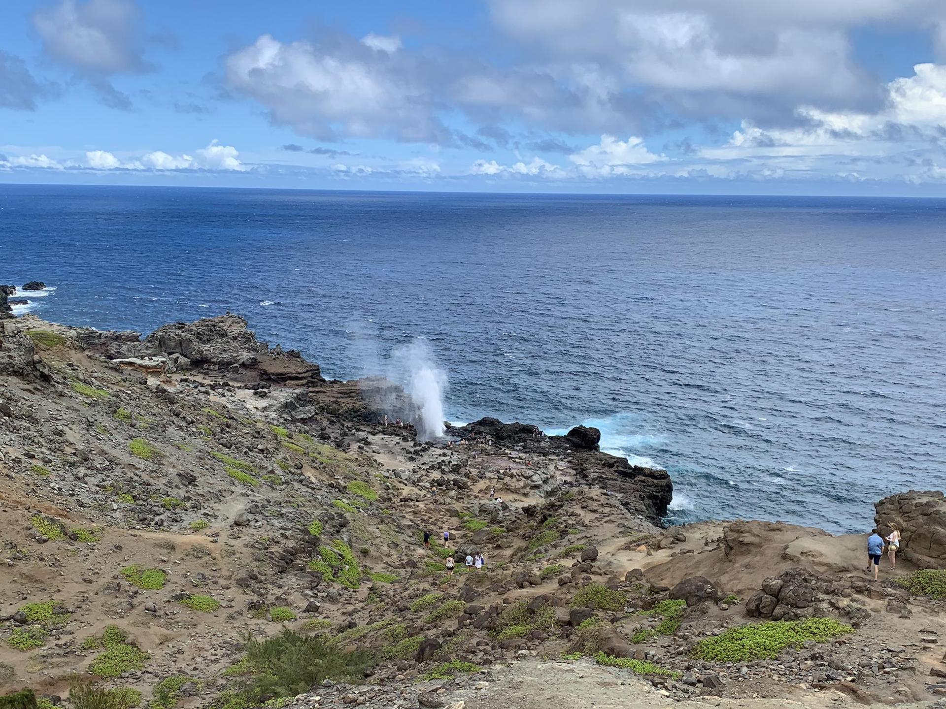 Coastal view of a rocky shore with a blowhole spraying water, people observing.