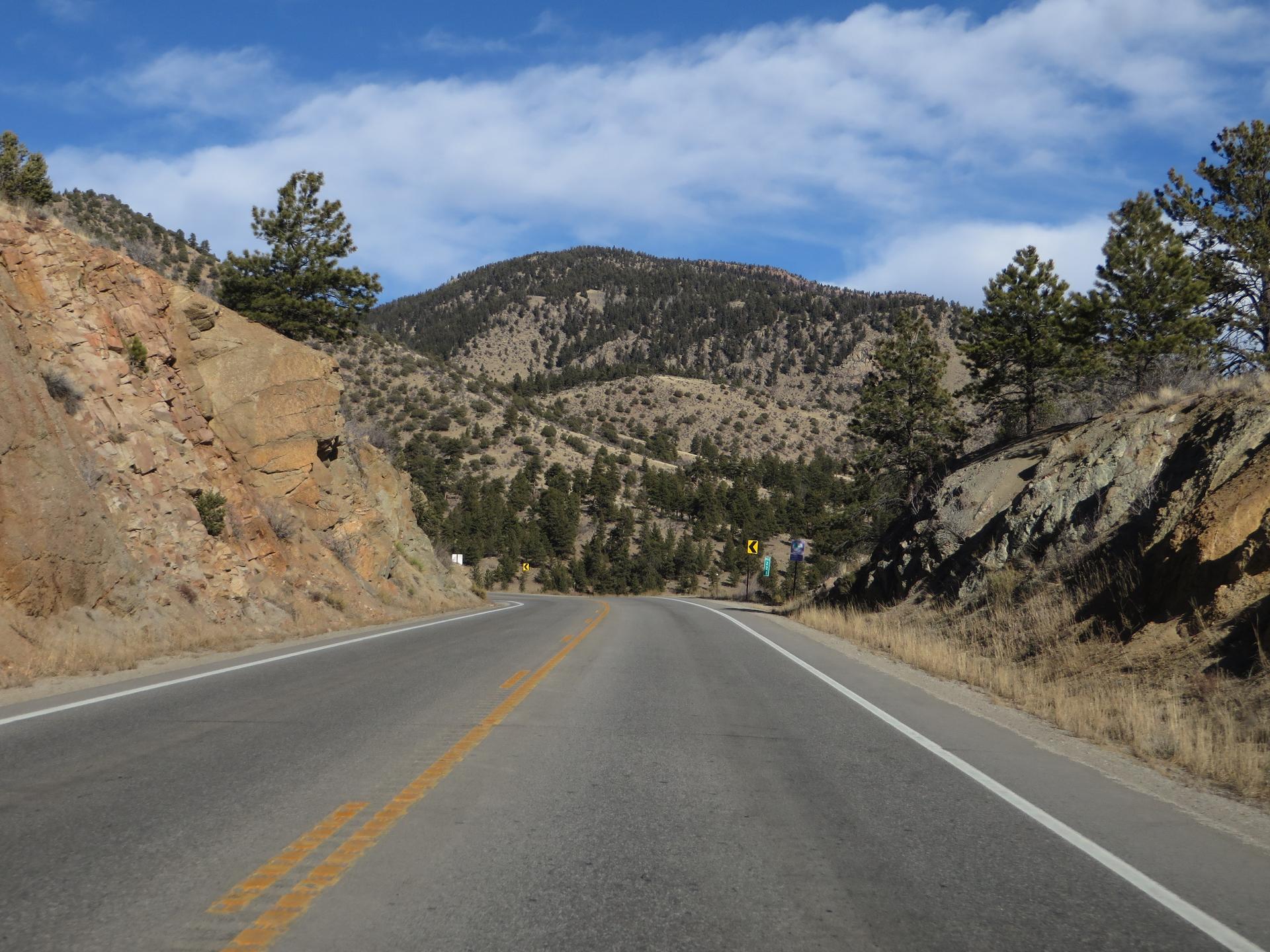 A winding road cuts through rocky, forested mountains under a bright, cloudy sky.
