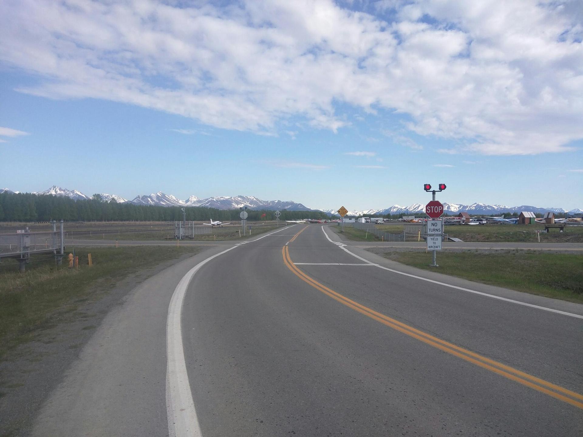 Paved road curving right towards distant snow-capped mountains under a bright, cloudy sky.
