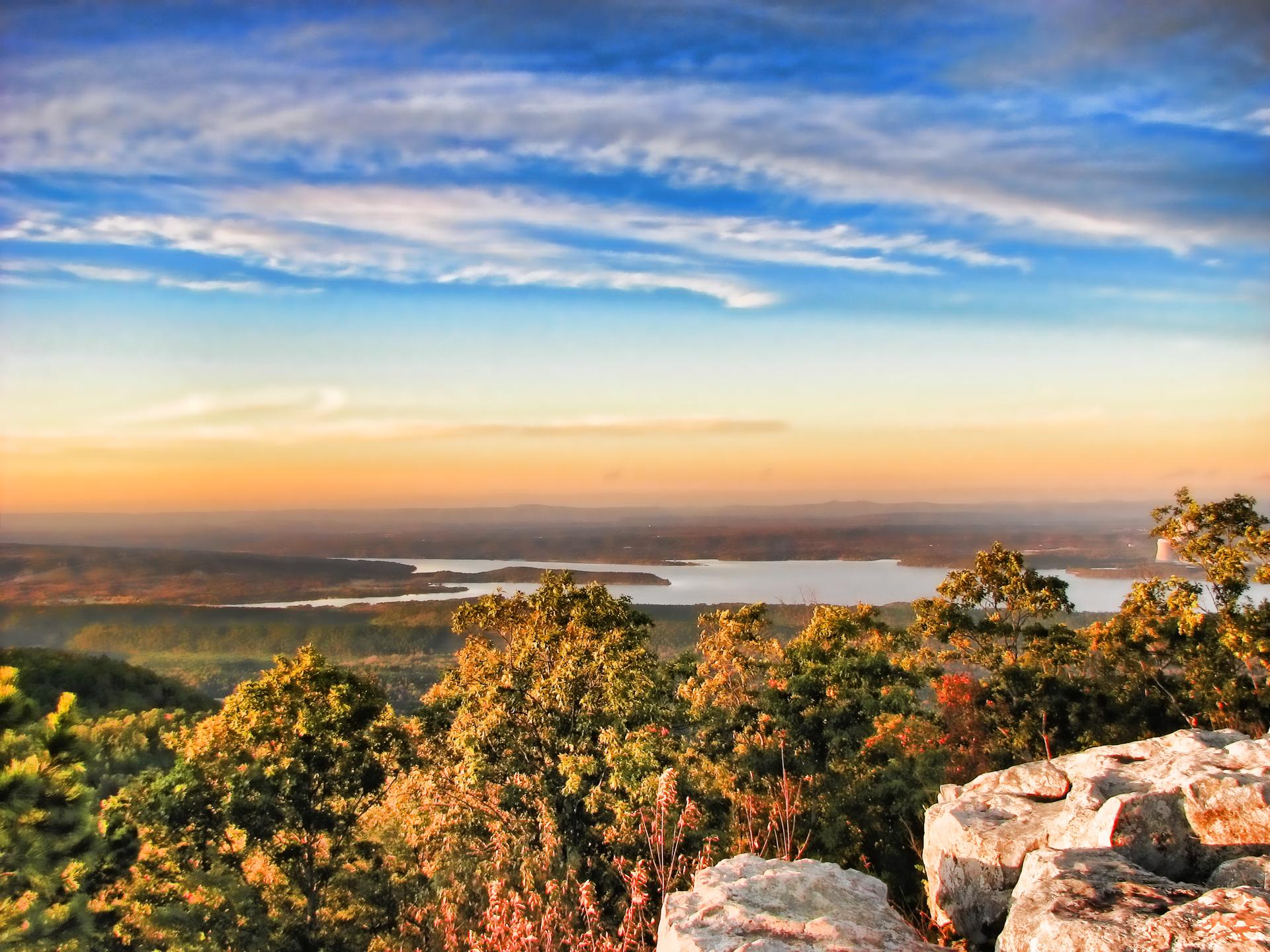 Vibrant sunset over a distant lake and forested hills from a rocky overlook.