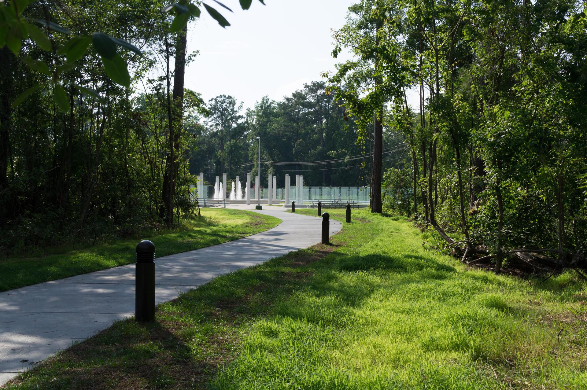 Winding paved path through woods towards white pillars in sunlight.