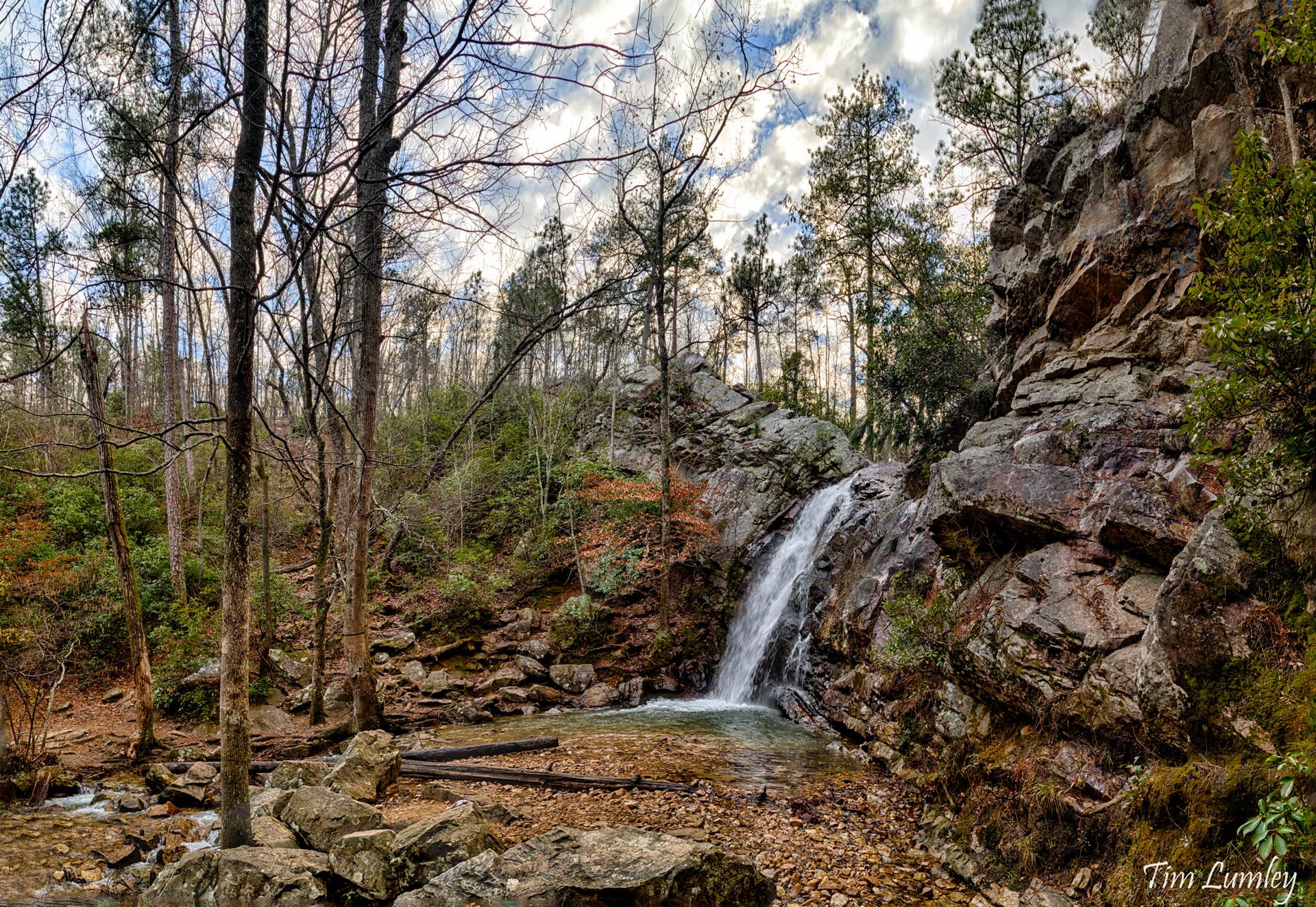 A waterfall cascades over rocky cliffs into a pool, surrounded by a sparse forest.