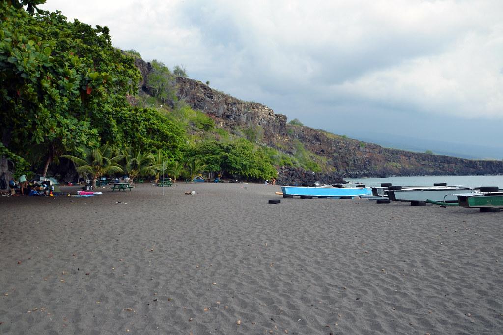 Black sand beach with boats, rocky cliff, and people under trees.