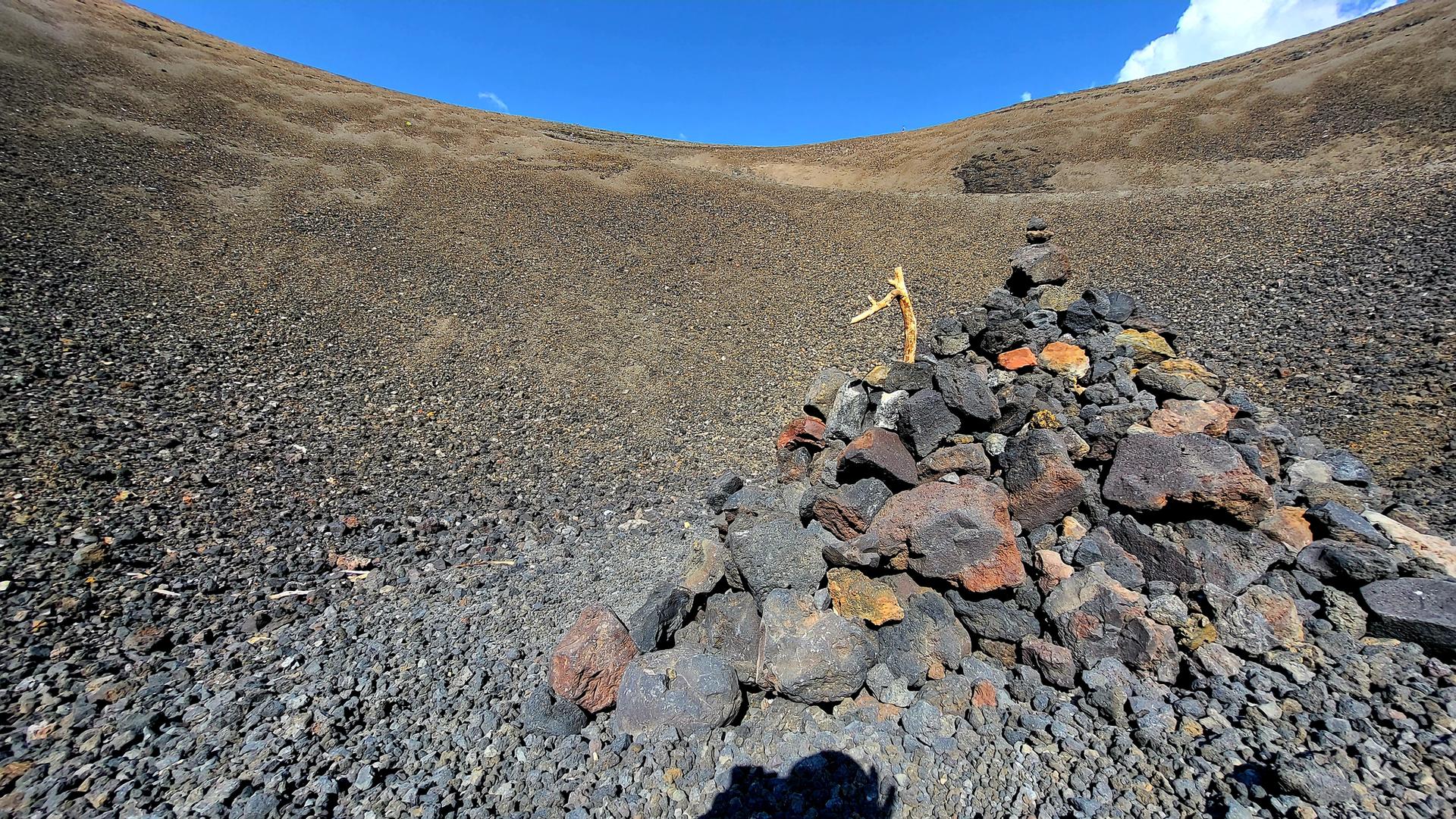 Close-up of a rock cairn inside a dark, rocky volcanic crater under a blue sky.