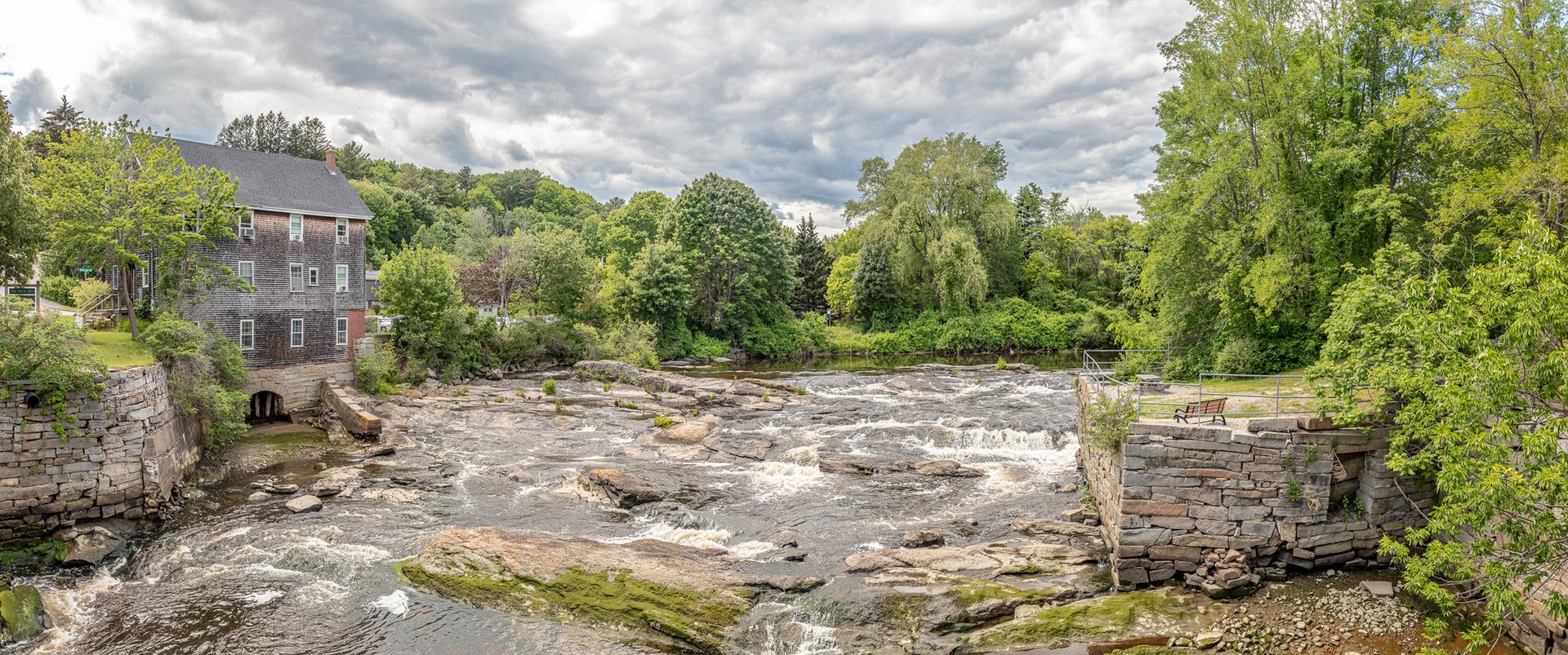 Panoramic view of a wide, rocky river with rapids and an old mill building.