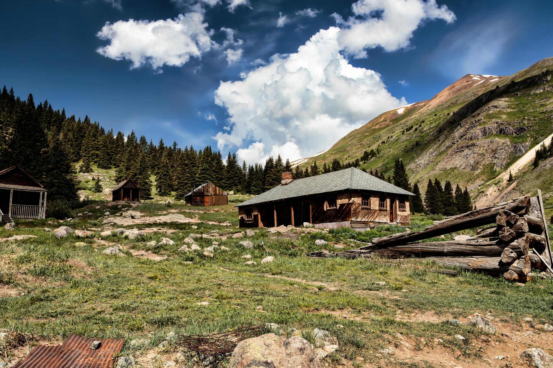 Historic wooden buildings on a grassy hillside with mountains and clouds.