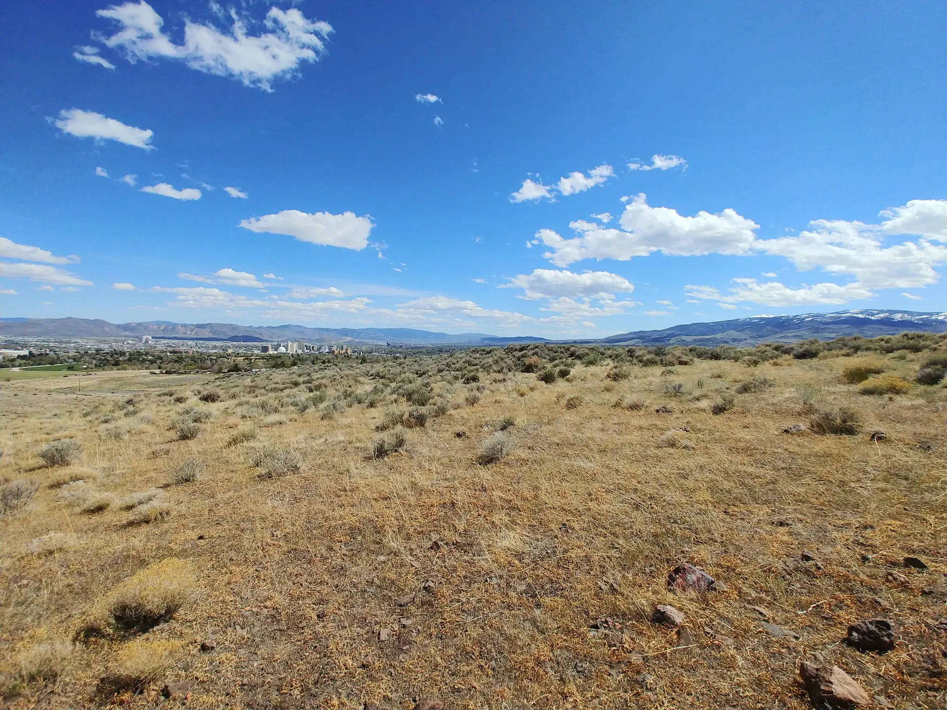 Wide-angle view of dry, brushy plains with a distant city under a bright blue sky.