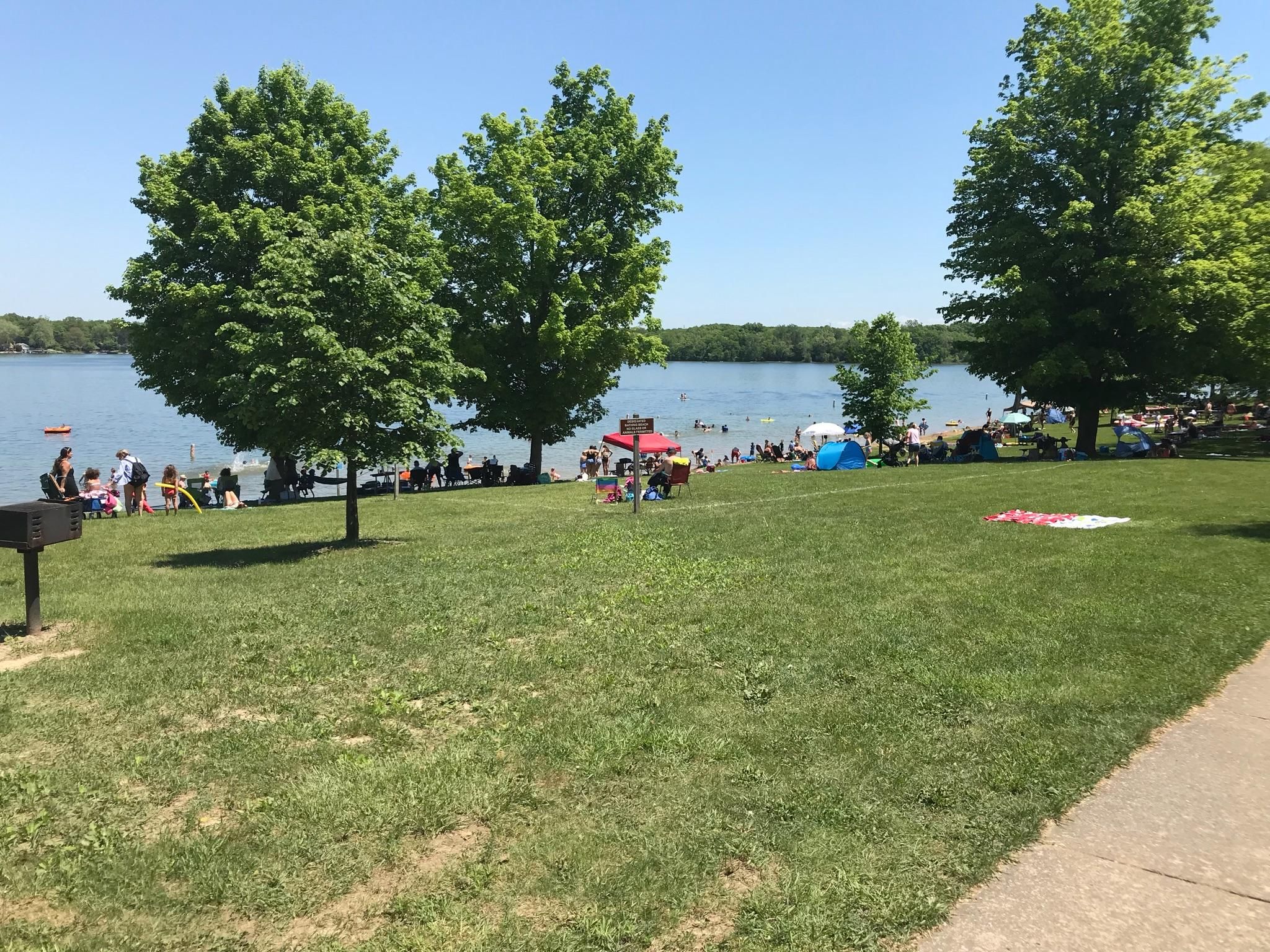 A grassy lawn with people relaxing by a lake, surrounded by trees. - Crooked Lake Trail