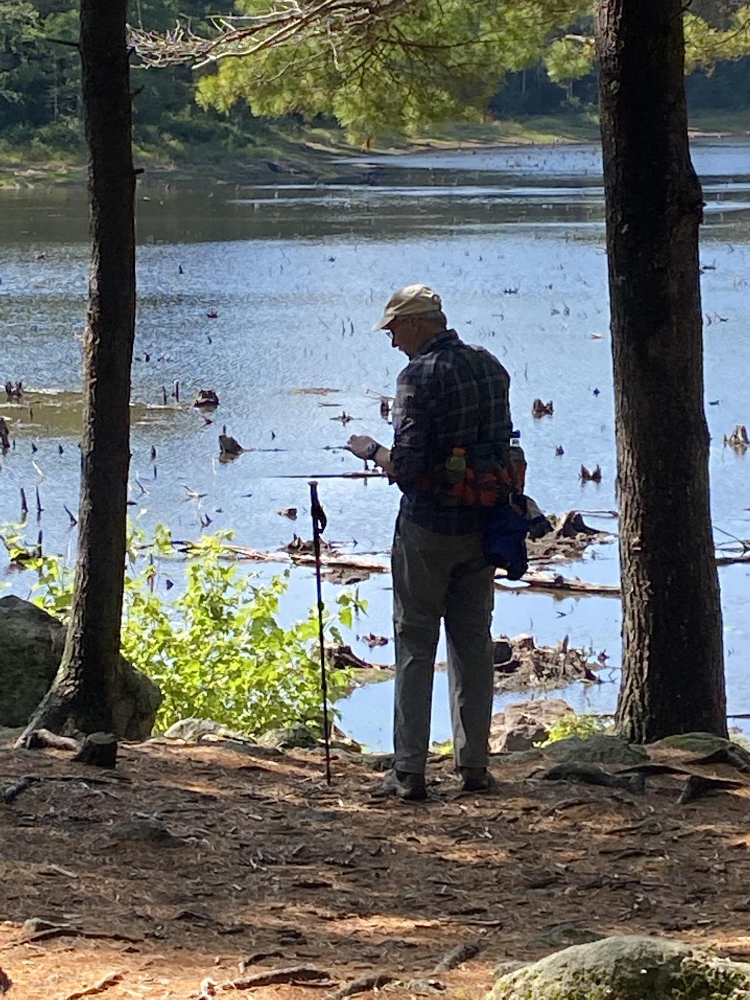 Man in plaid shirt looking at a map by a lake, framed by trees.