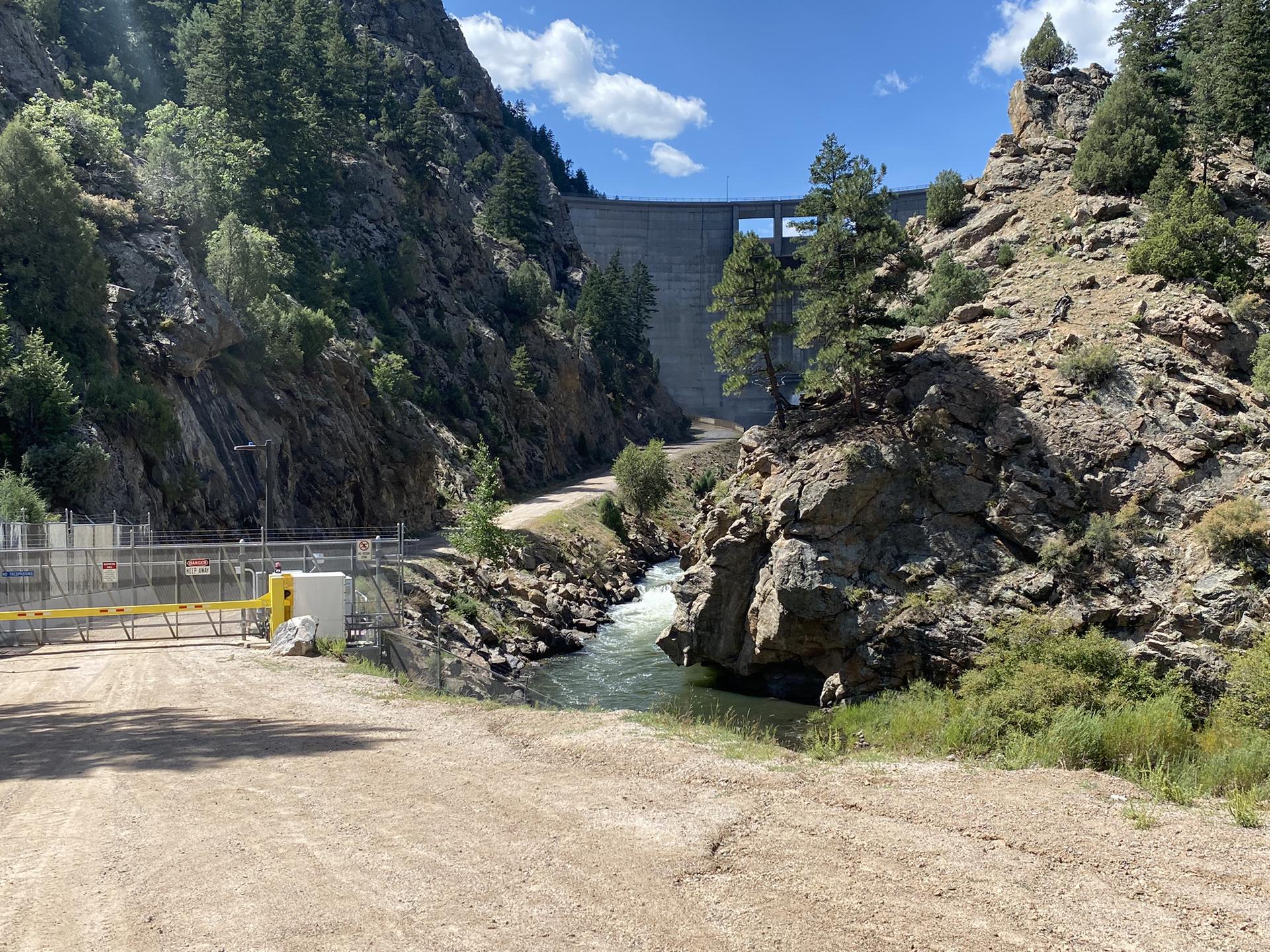 Large dam in a rocky canyon with a river and dirt road on a sunny day.