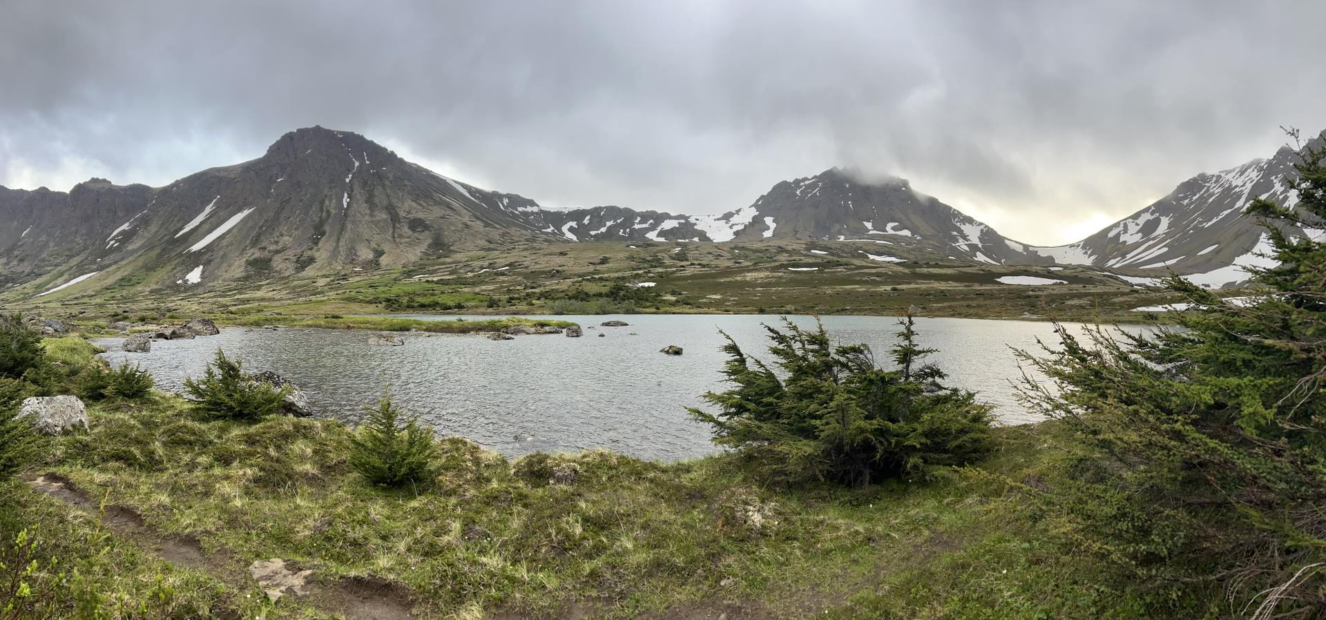Panoramic view of a lake surrounded by snowy mountains under a cloudy sky.