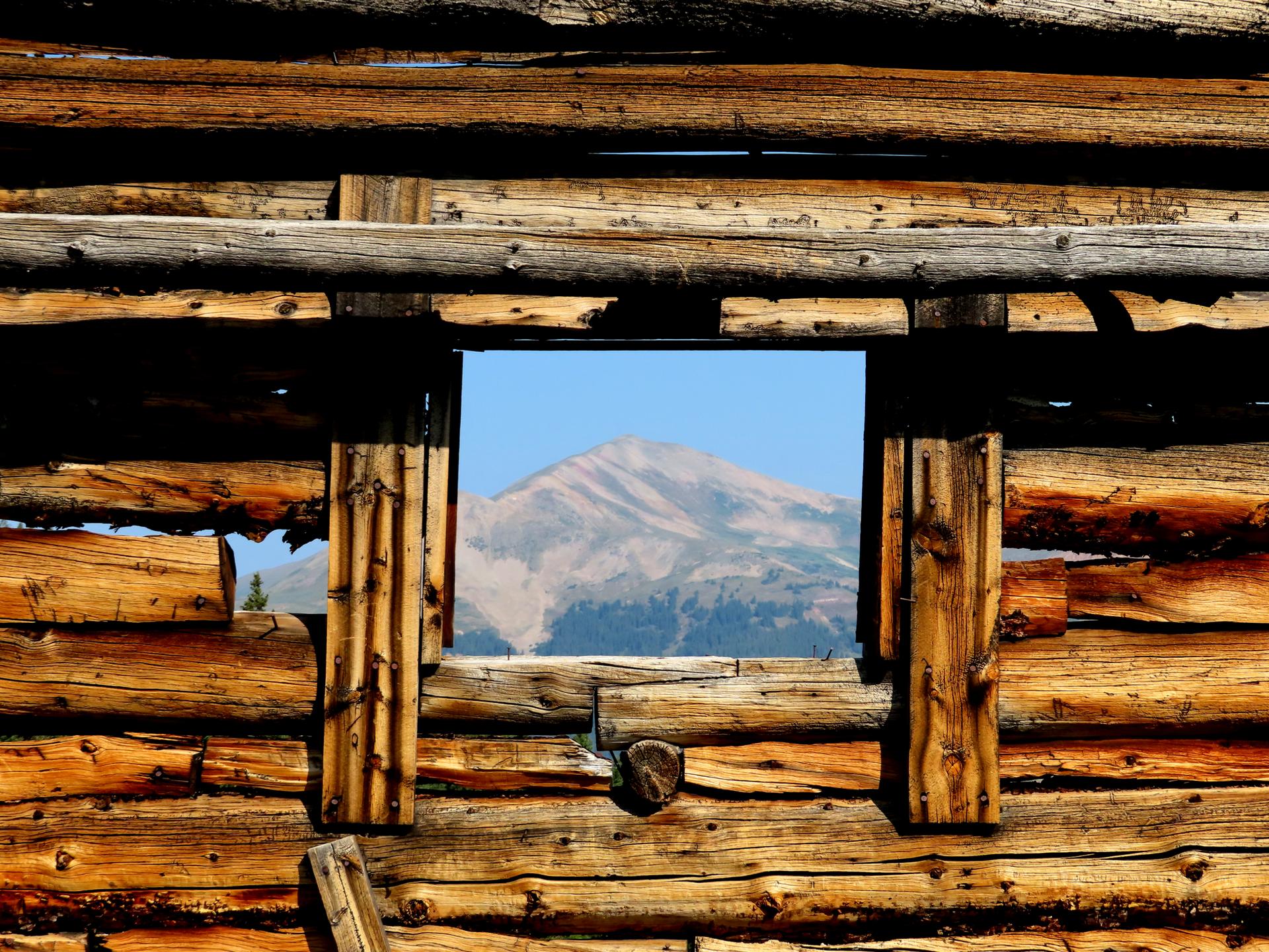 Mountain peak seen through a rustic log cabin window on a sunny day.