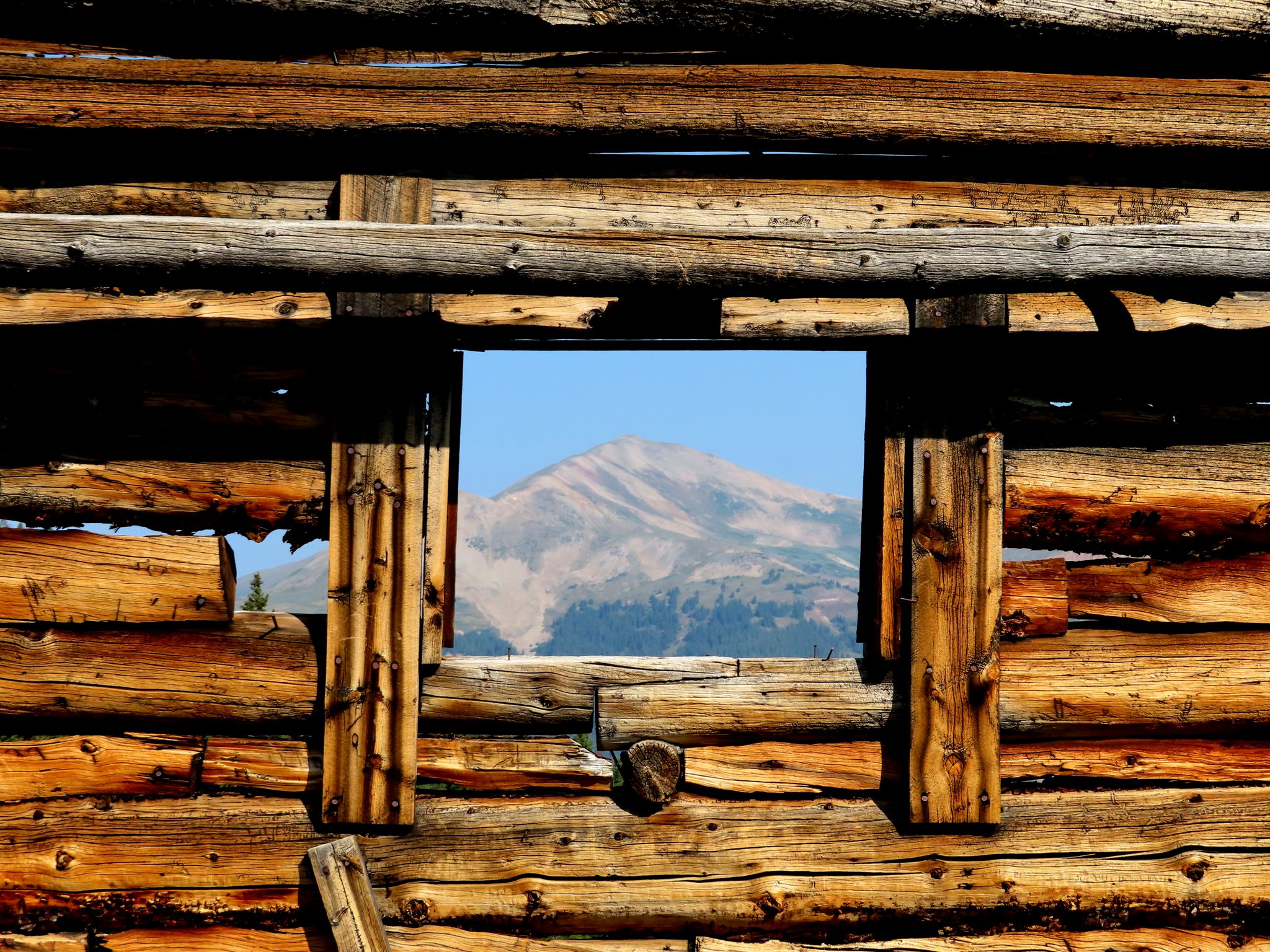 Mountain peak seen through a rustic log cabin window on a sunny day. - Mayflower Gulch Trail