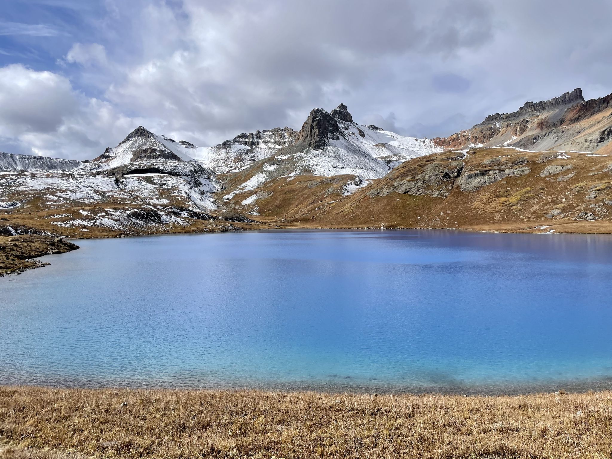 Bright blue alpine lake surrounded by snowy, rugged mountains under clouds. - Ice Lakes Trail