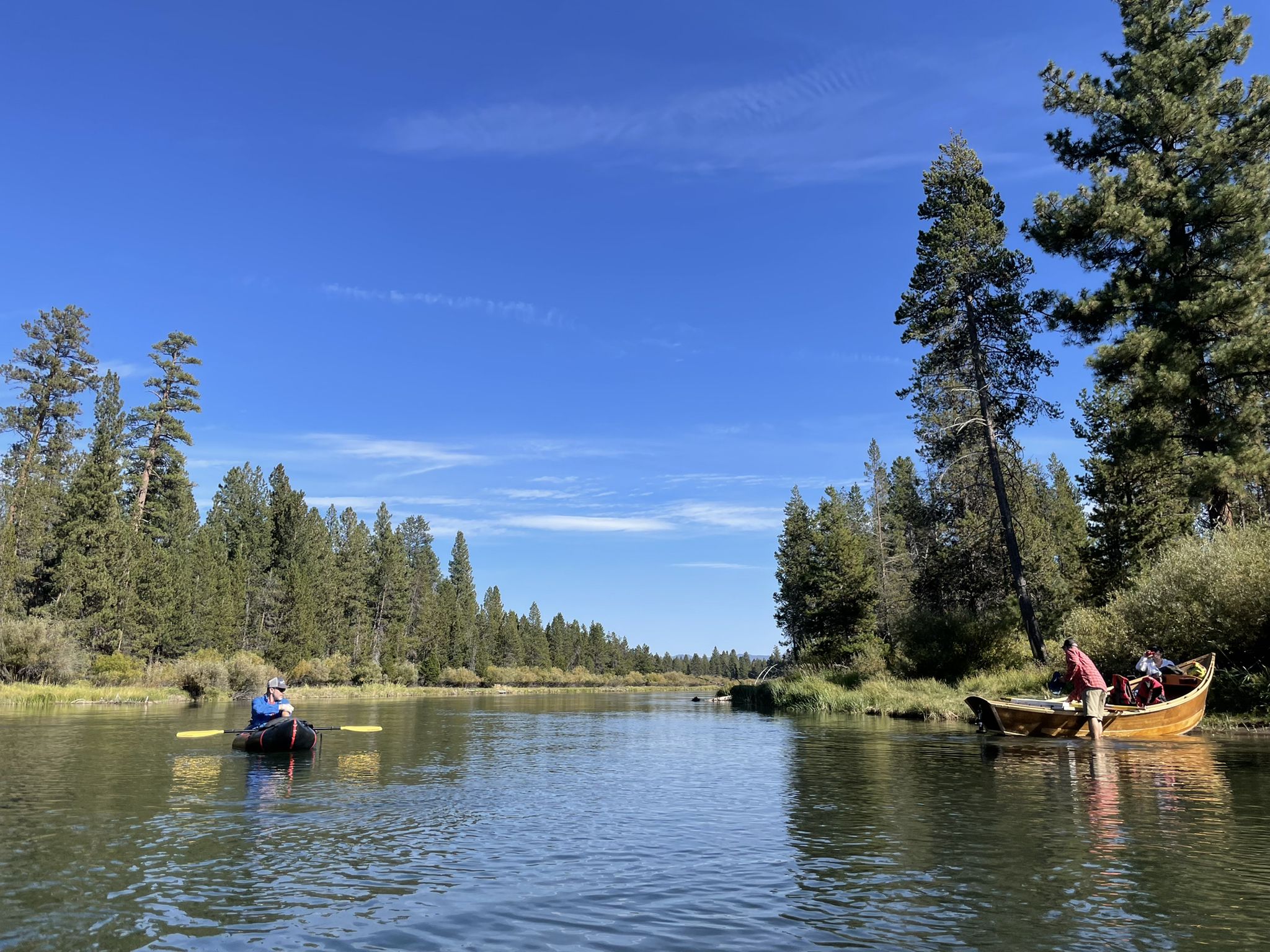 Two people paddle boats on a sunny river lined with tall pine trees. - Big River Campground loop from La Pine State Park