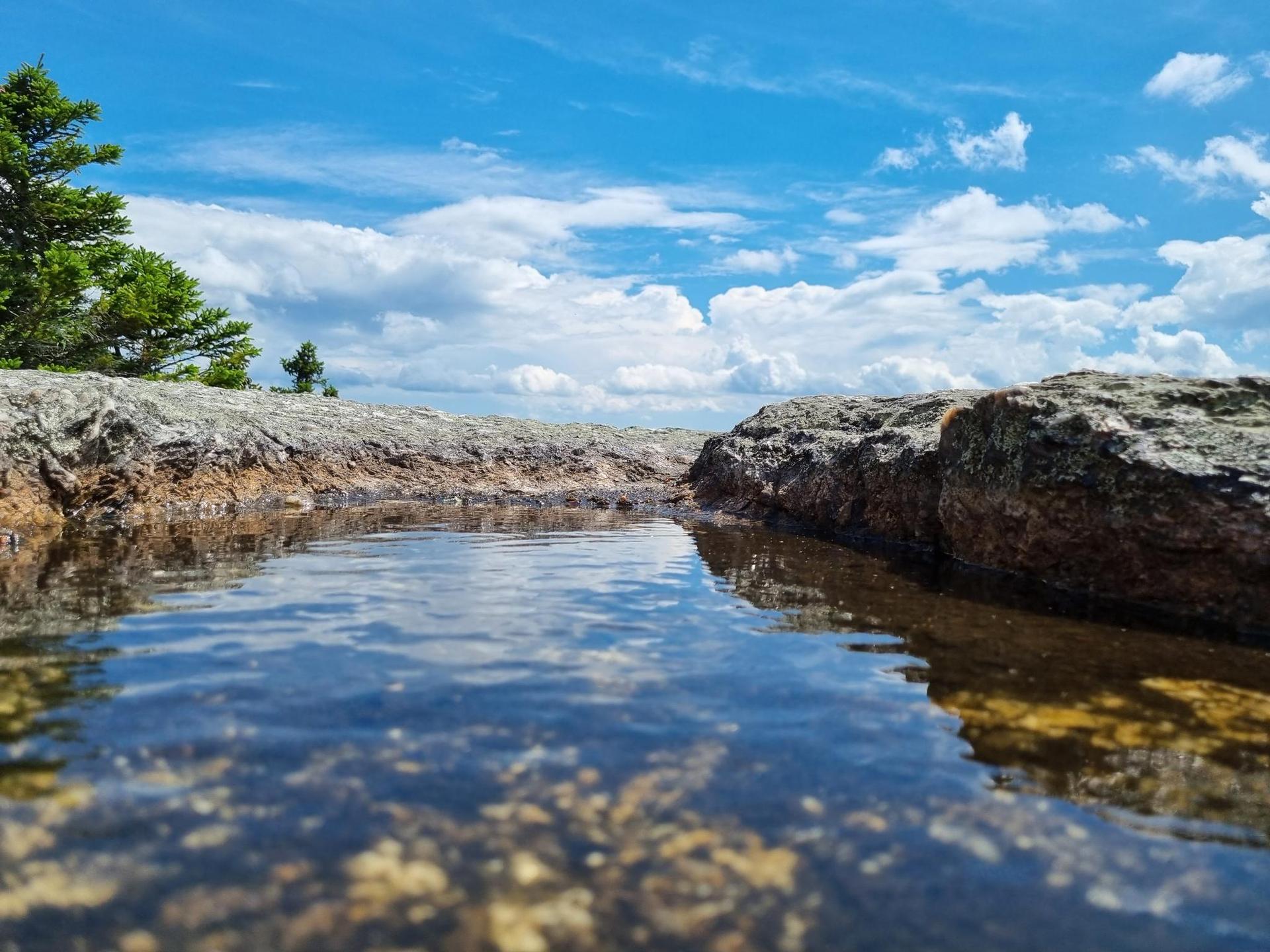 Shallow water reflecting a bright blue sky with clouds over rocky terrain.