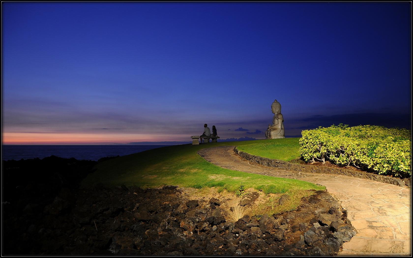 Two people sit on a bench overlooking the ocean at twilight.