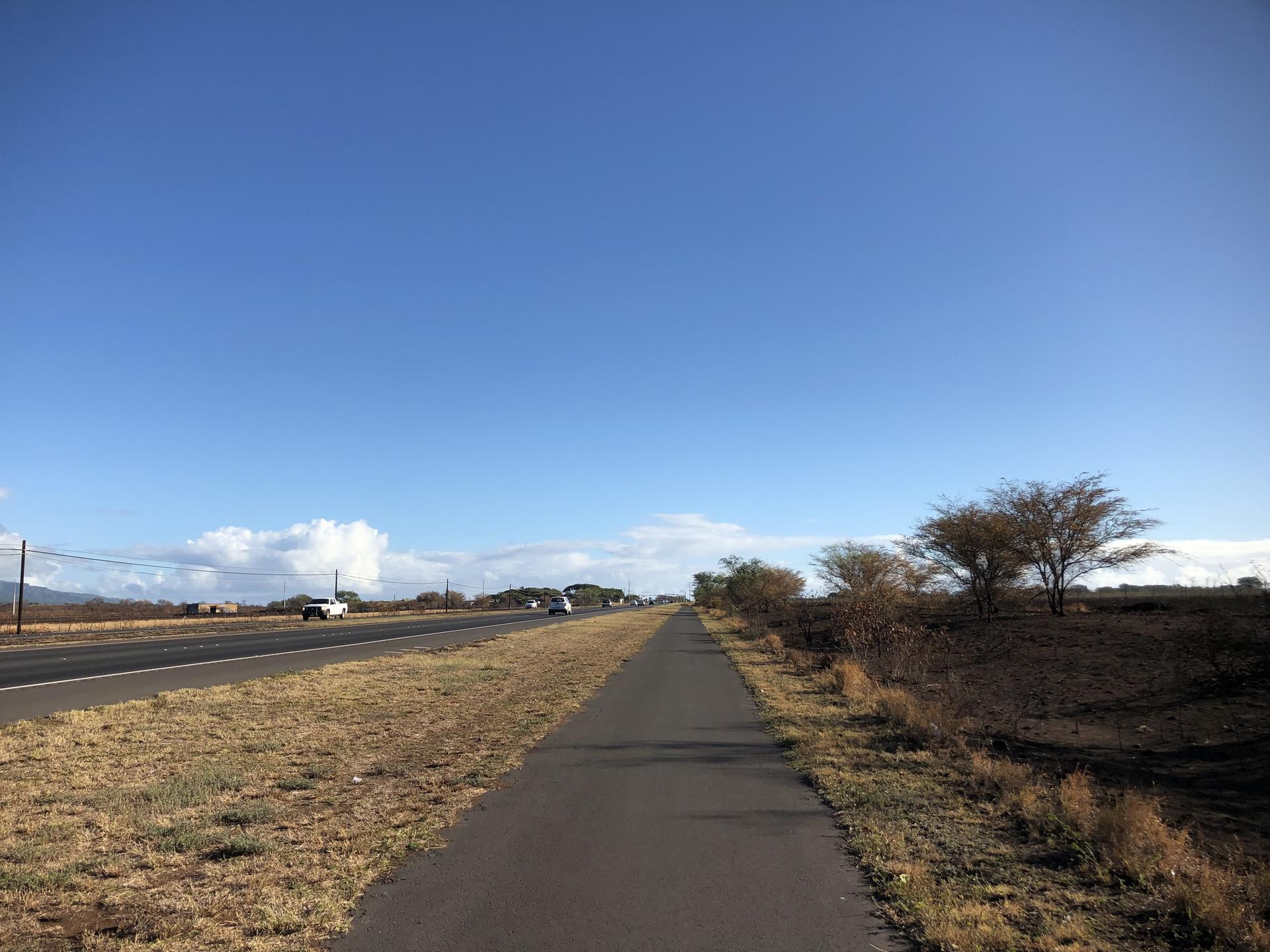 Sunny day with a road and parallel path through dry, open landscape.