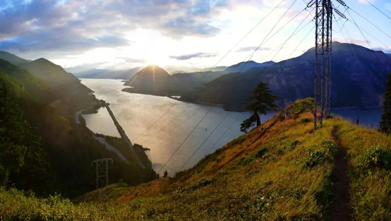 Golden sunset over a wide river, mountains, power lines, and hikers on a path.