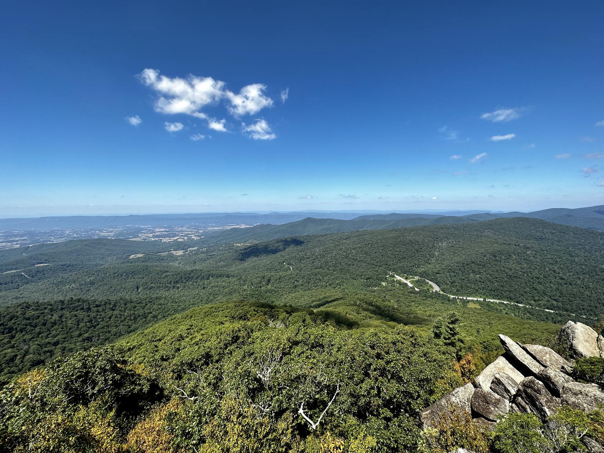 Panoramic view of green forested mountains and a winding road under a blue sky.