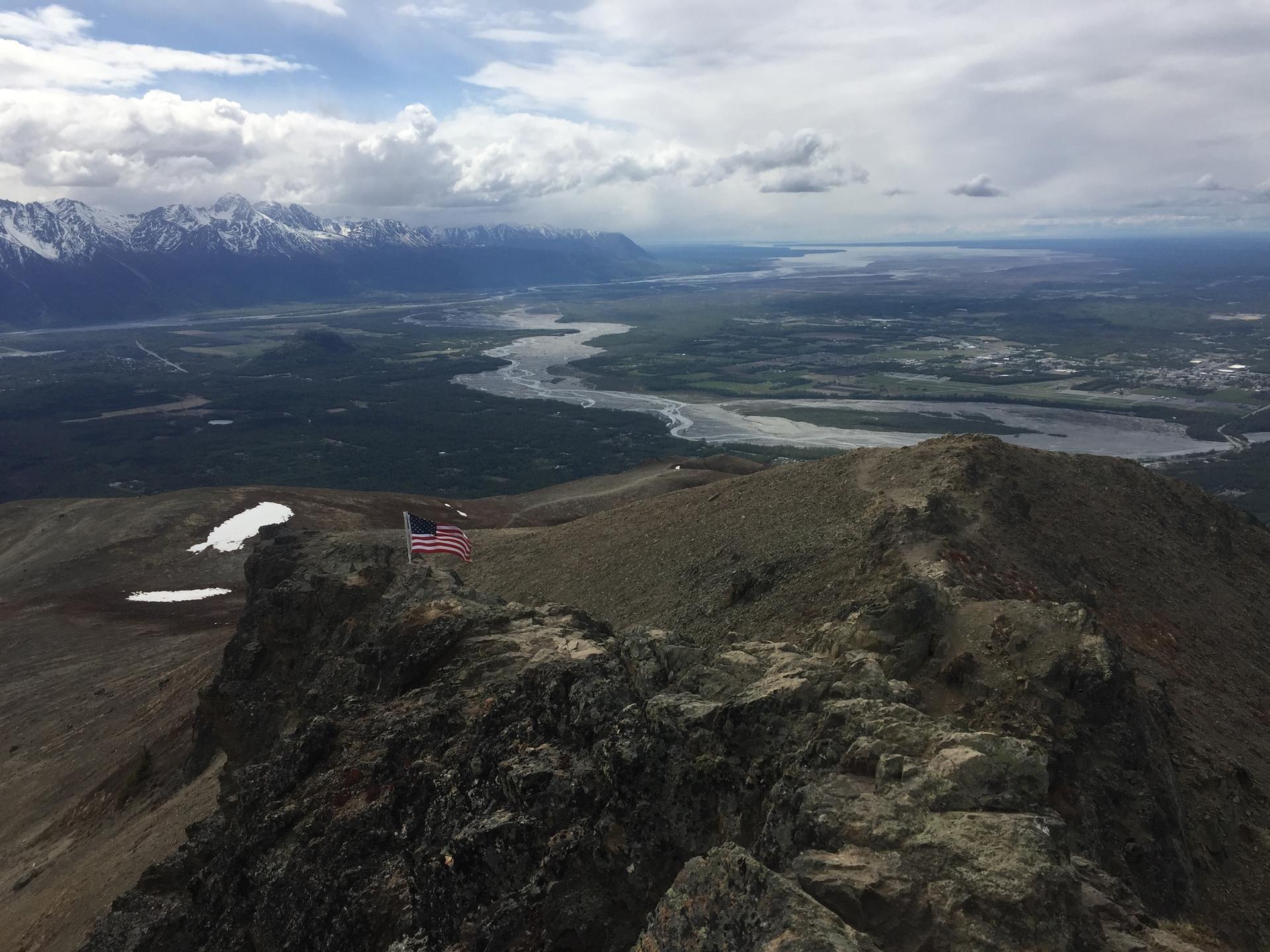 Rocky mountain ridge with a flag, overlooking a vast river valley and distant peaks.