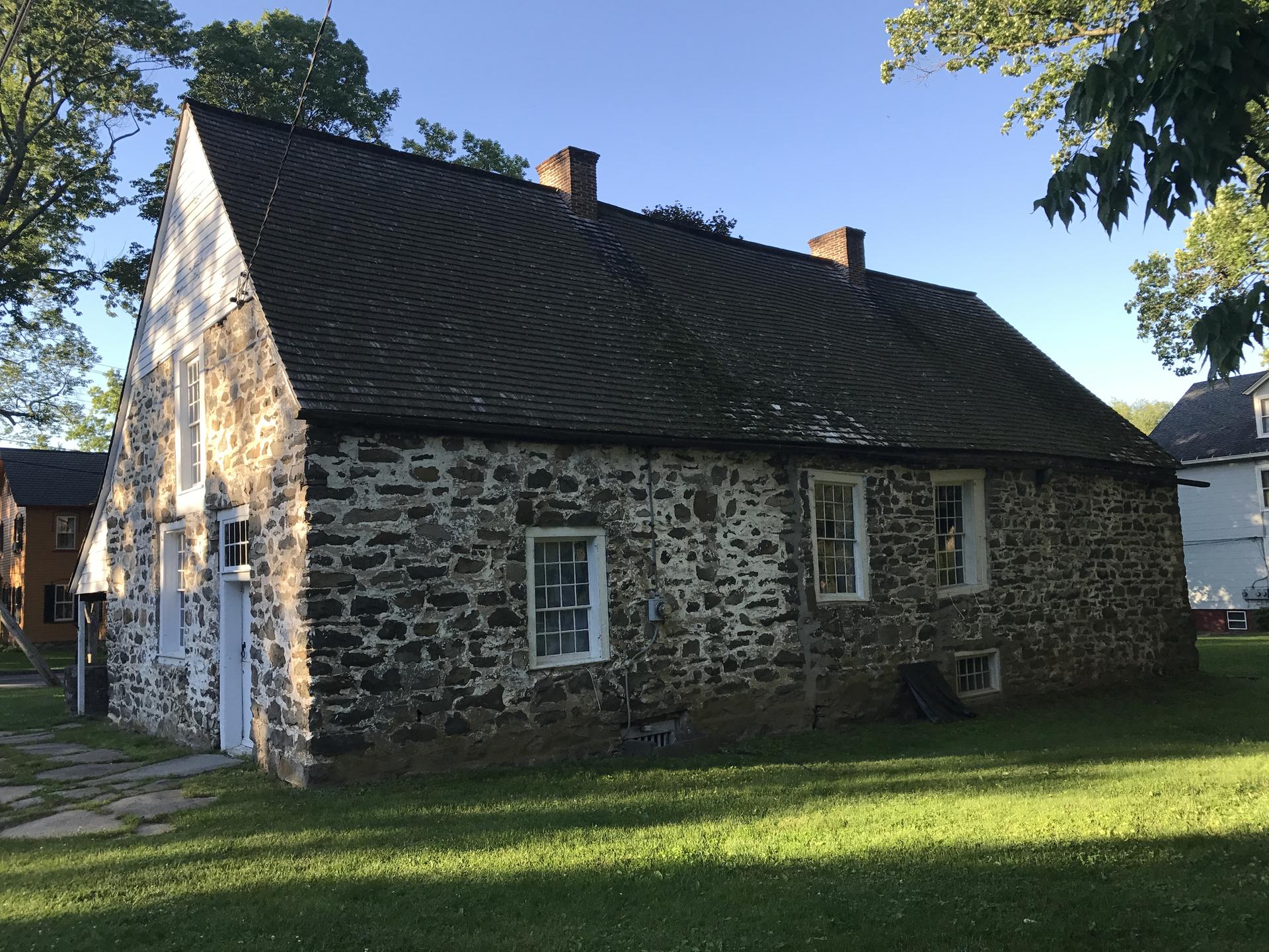 Historic stone house with a dark roof and white siding under bright sunlight.