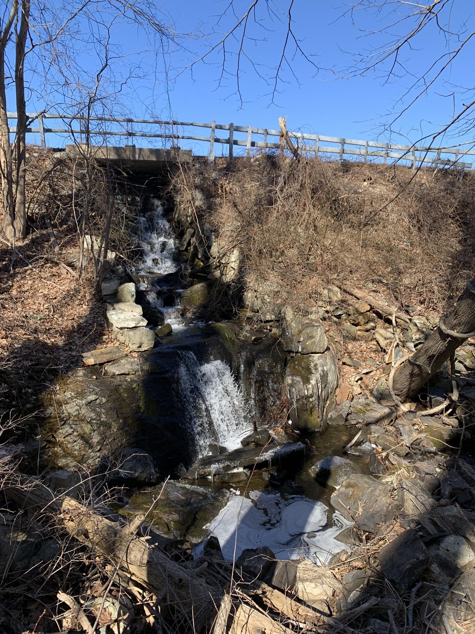 Small waterfall cascading over rocks under a bridge on a sunny day.