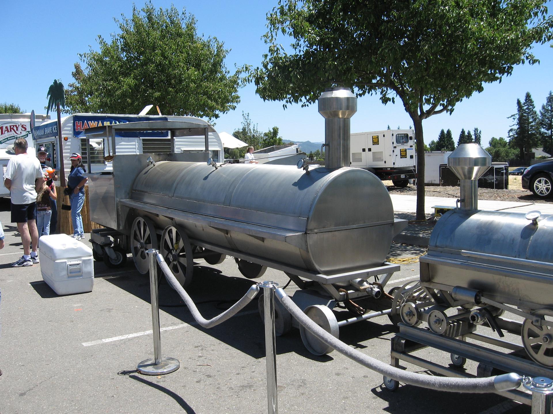 Large train-shaped barbecue smokers outdoors with people under bright daylight.