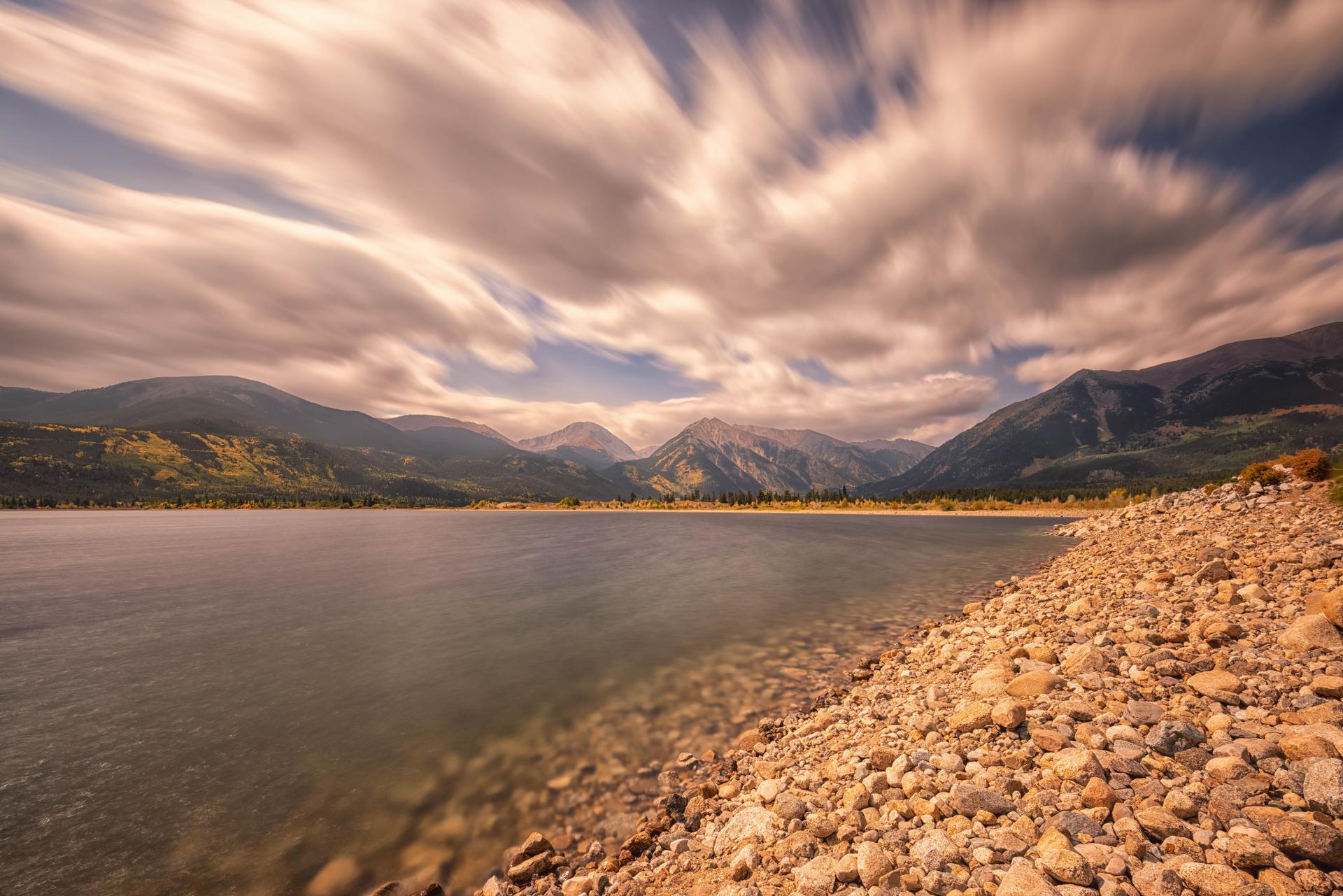 A serene lake with a rocky shore and mountains under a cloudy sky.
