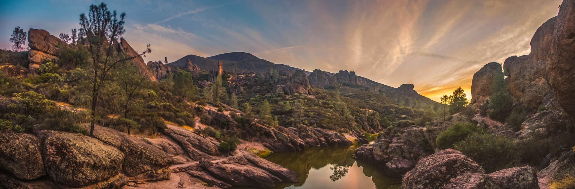 Panoramic sunset over a calm lake surrounded by rugged rocky mountains and sparse trees.
