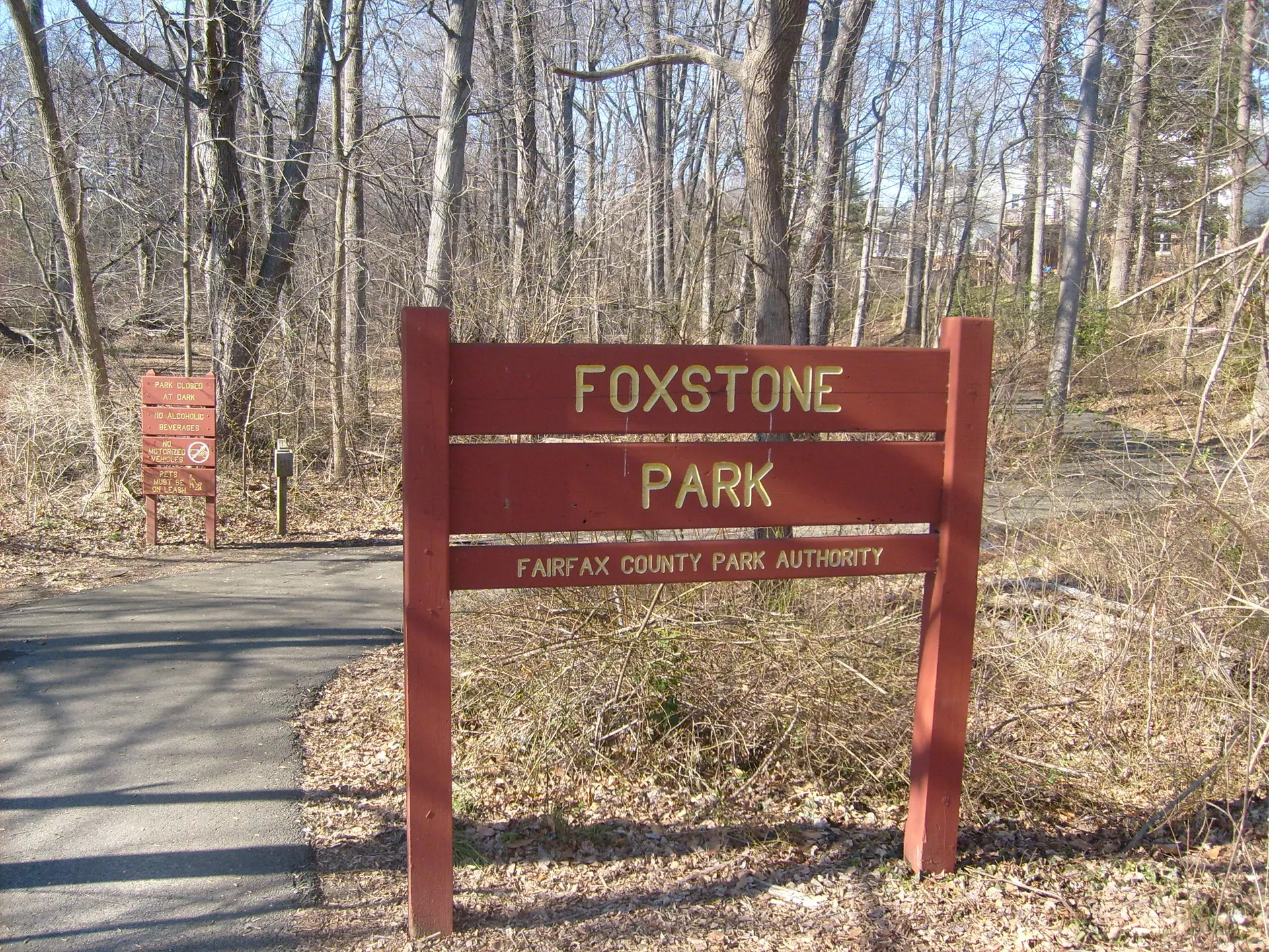 A wooden sign stands beside a paved path leading into a bare winter forest.