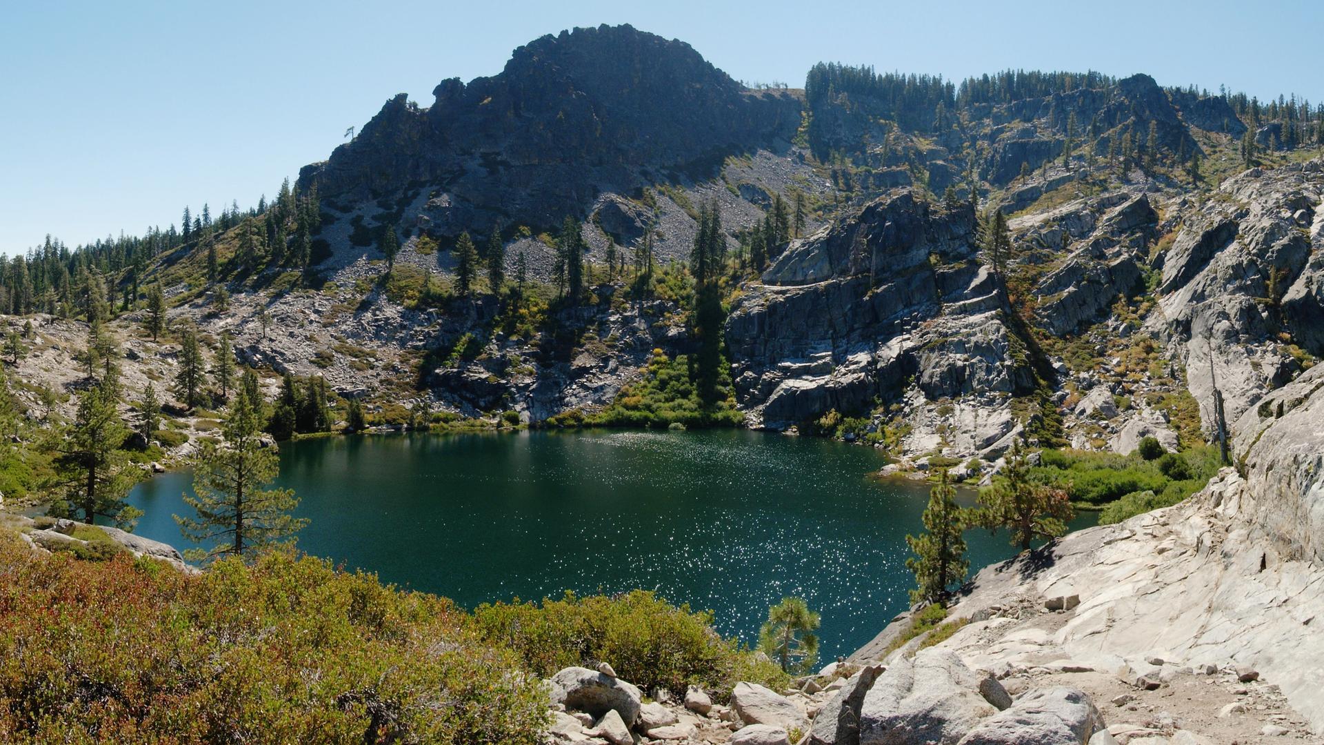 Dark green mountain lake nestled among rugged rocky peaks and sparse trees.