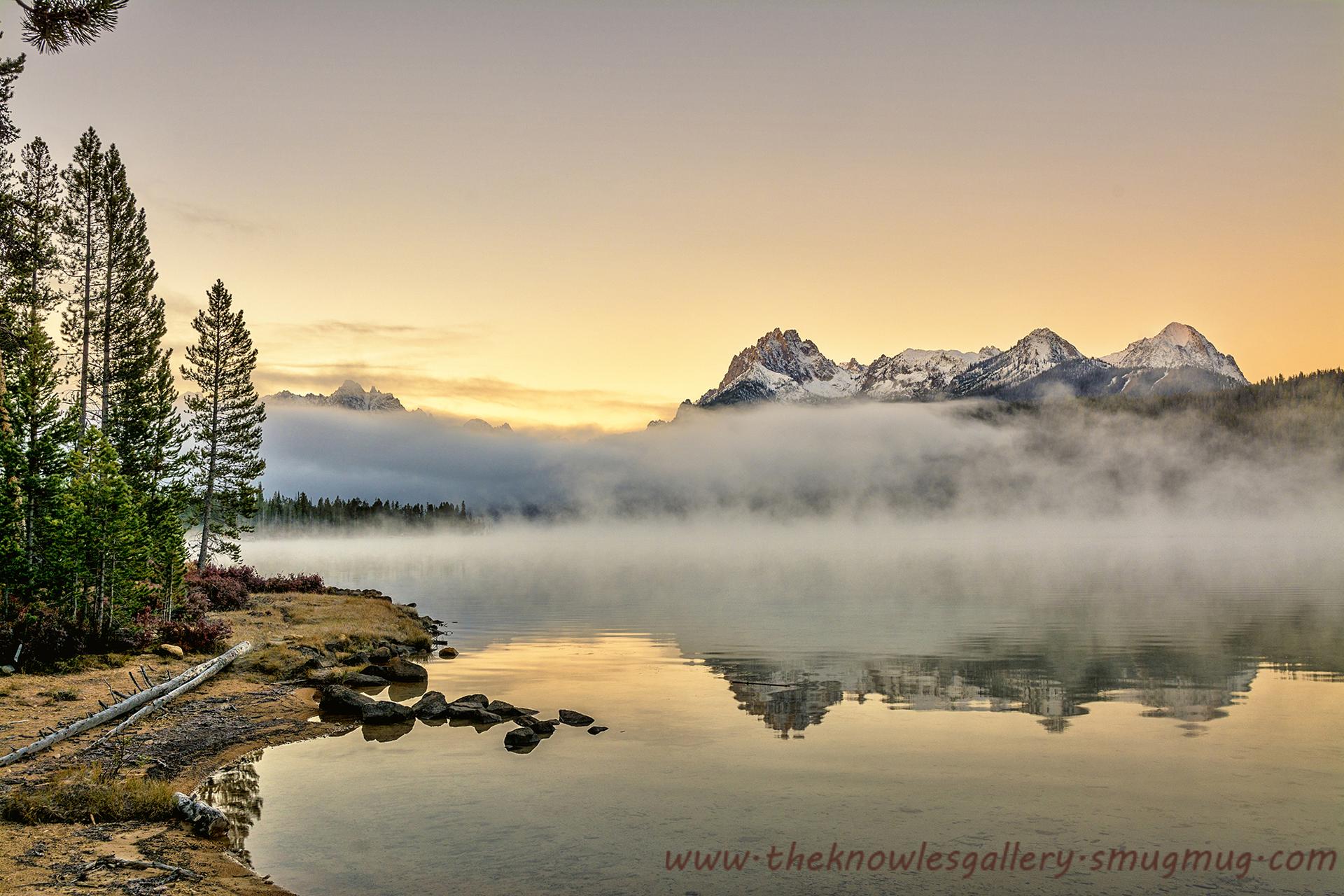 Misty lake at sunrise with pine trees and snow-capped mountains reflecting in water.
