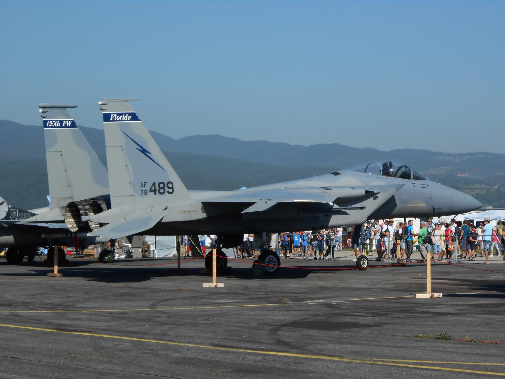 Grey fighter jet on an airfield with mountains, people viewing under bright sun.