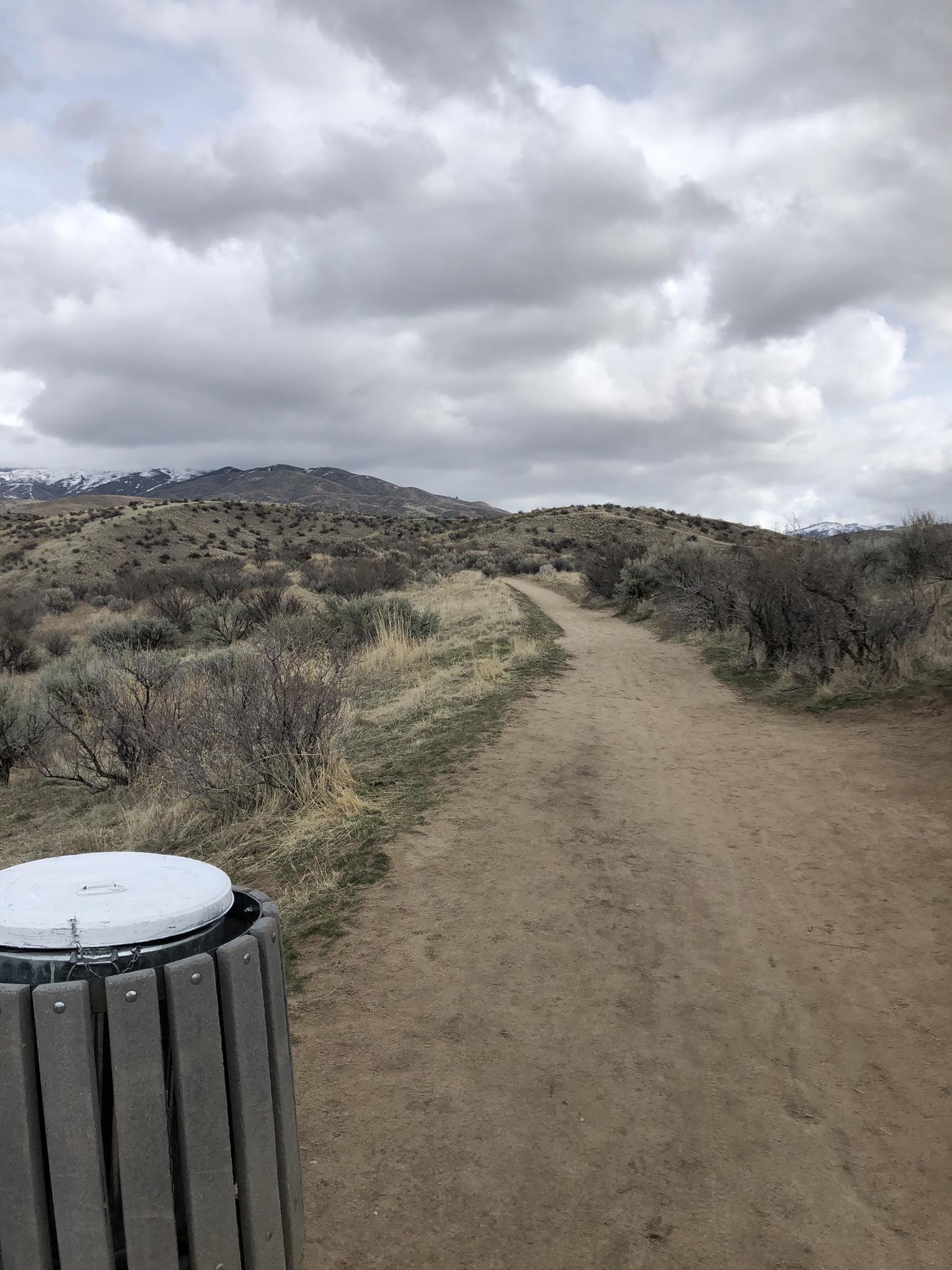 A wide dirt path next to a trash can, leading towards distant hills under an overcast sky.