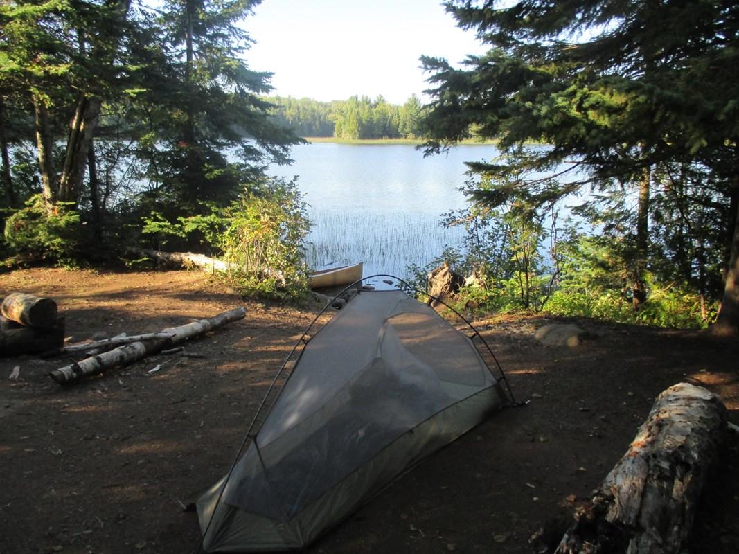 A tent pitched on a sunny forested shore overlooking a calm lake with a canoe.