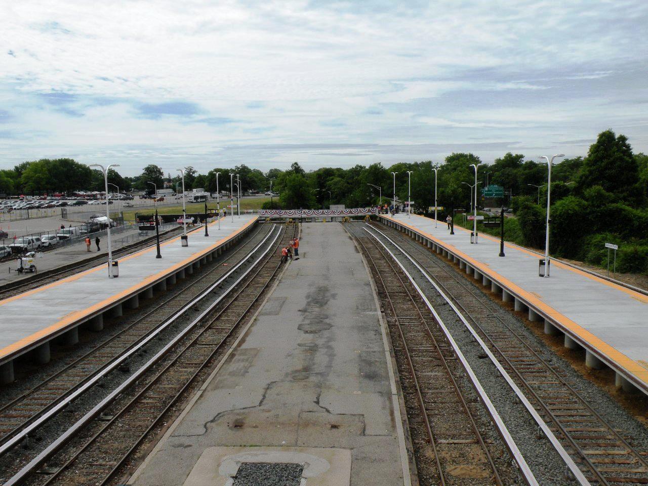 Train station platforms and tracks with workers under a bright, cloudy sky.